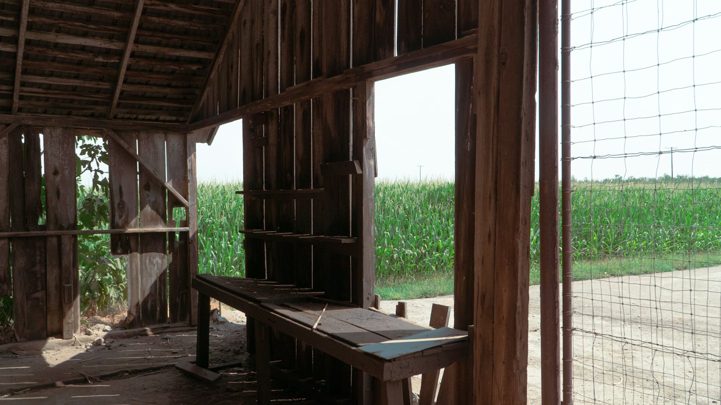A weathered wooden workbench sits inside a rustic barn overlooking green cornfields.